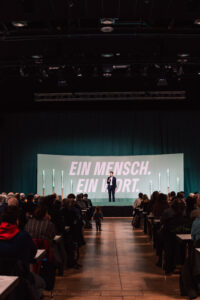 Max Lucks, Abgeordneter der Grünen, steht auf einer großen Bühne bei einer Wahlkampfveranstaltung. Im Vordergrund sitzen die Zuschauer*innen, ein Kind läuft über den Gang.