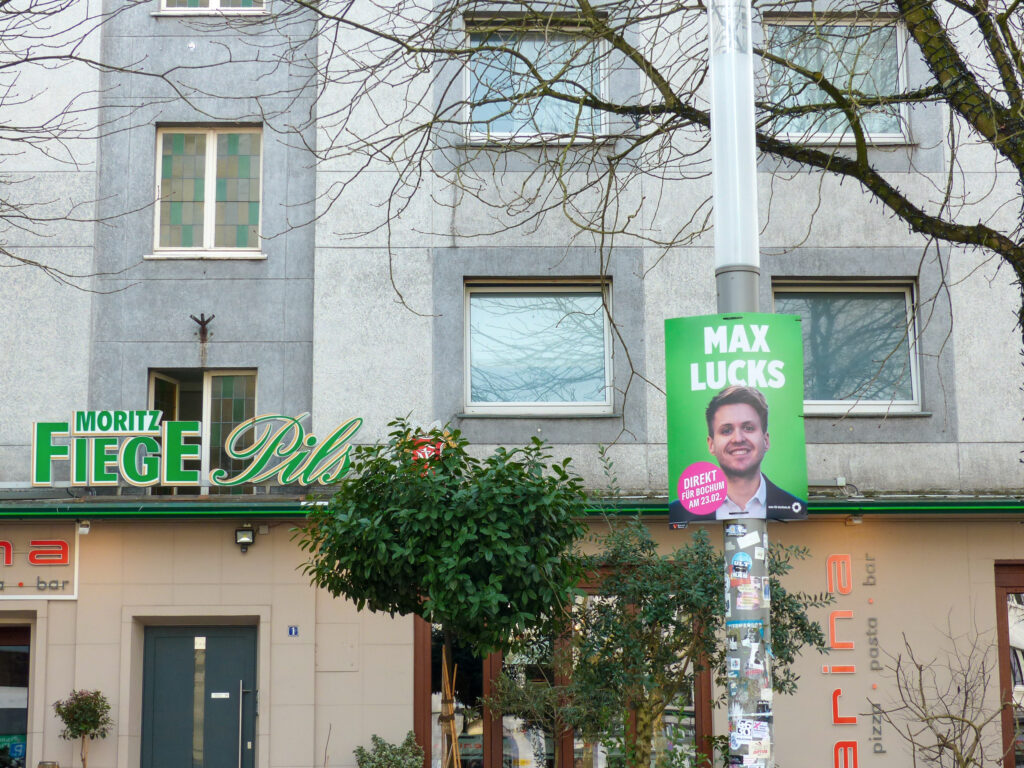 Ein Wahlplakat des Grünen-Politikers Max Lucks häng vor einem Moritz Fiege Schild, das an einem Gebäude befestigt ist. 