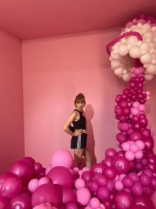 Carla steht in einer Menge pinker Luftballons die von rechts oben nach links unten drapiert sind.