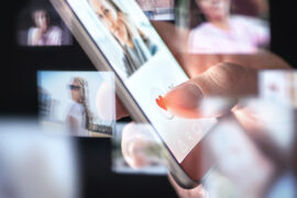 Handy in der Hand eines Menschen. Darauf ist ein Dating-Profil geöffnet. Drum herum schwirren noch etliche andere Fotos von Profilen.