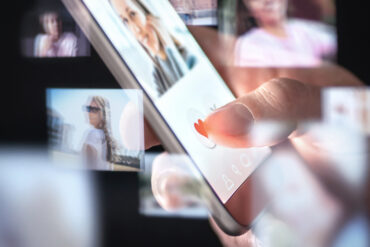 Handy in der Hand eines Menschen. Darauf ist ein Dating-Profil geöffnet. Drum herum schwirren noch etliche andere Fotos von Profilen.