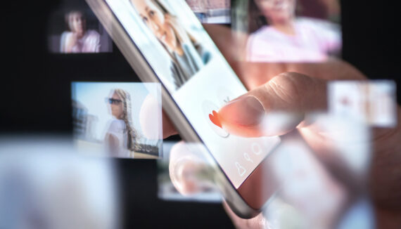 Handy in der Hand eines Menschen. Darauf ist ein Dating-Profil geöffnet. Drum herum schwirren noch etliche andere Fotos von Profilen.