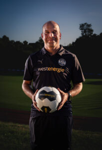 Christian Kowalski, Koordinator Mädchenfußball der SGS Essen, steht mit einem Ball vor einem Fußballfeld.