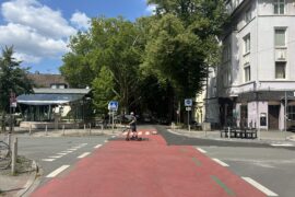 Die Große Heimstraße in Dortmund auf höhe der Stadtbahn-Haltestelle Kreuzstraße. Das Bild zeigt einen E-Scooter Fahrer der die rote Fahrbahn überquert.