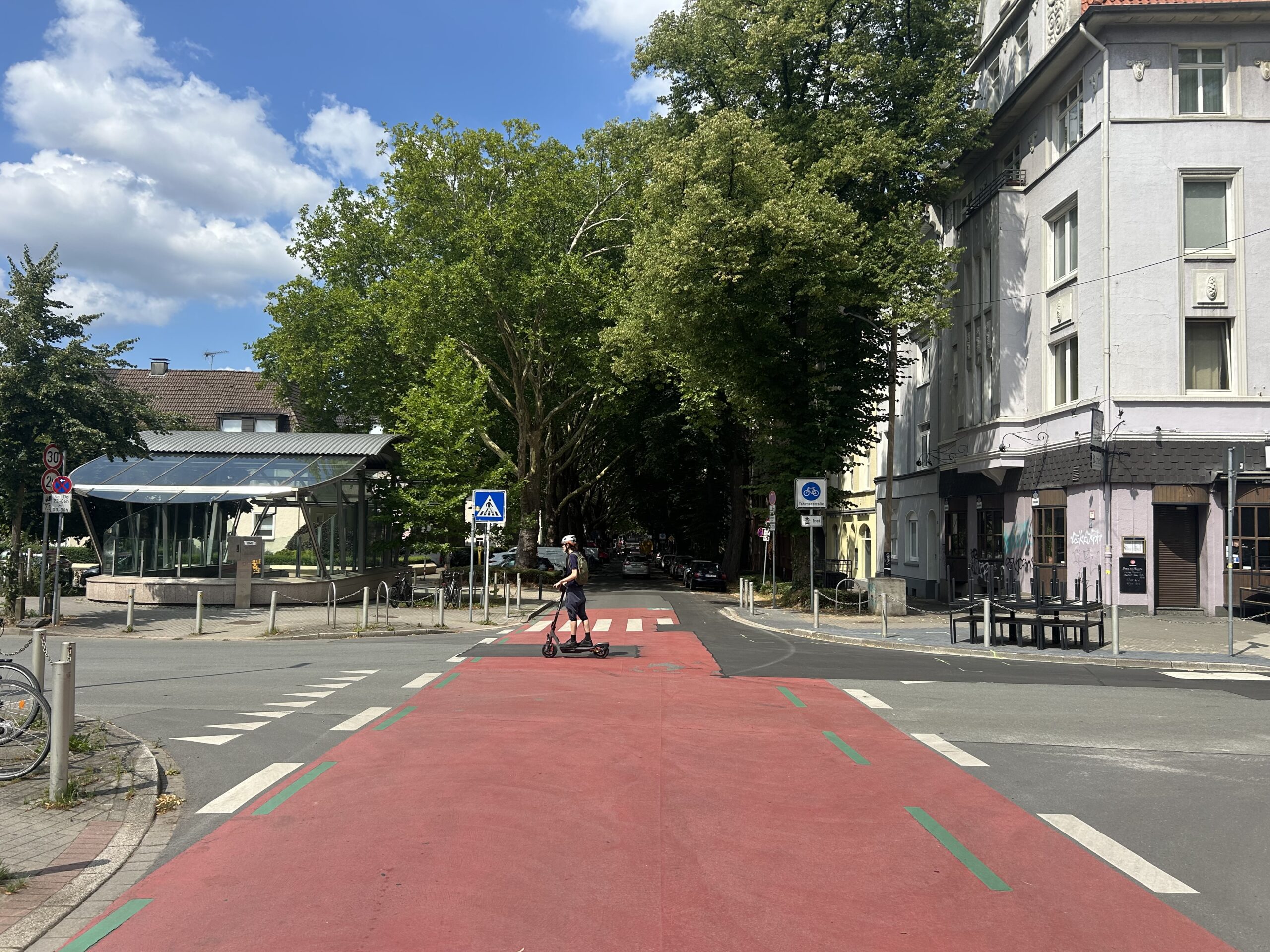 Die Große Heimstraße in Dortmund auf höhe der Stadtbahn-Haltestelle Kreuzstraße. Das Bild zeigt einen E-Scooter Fahrer der die rote Fahrbahn überquert.