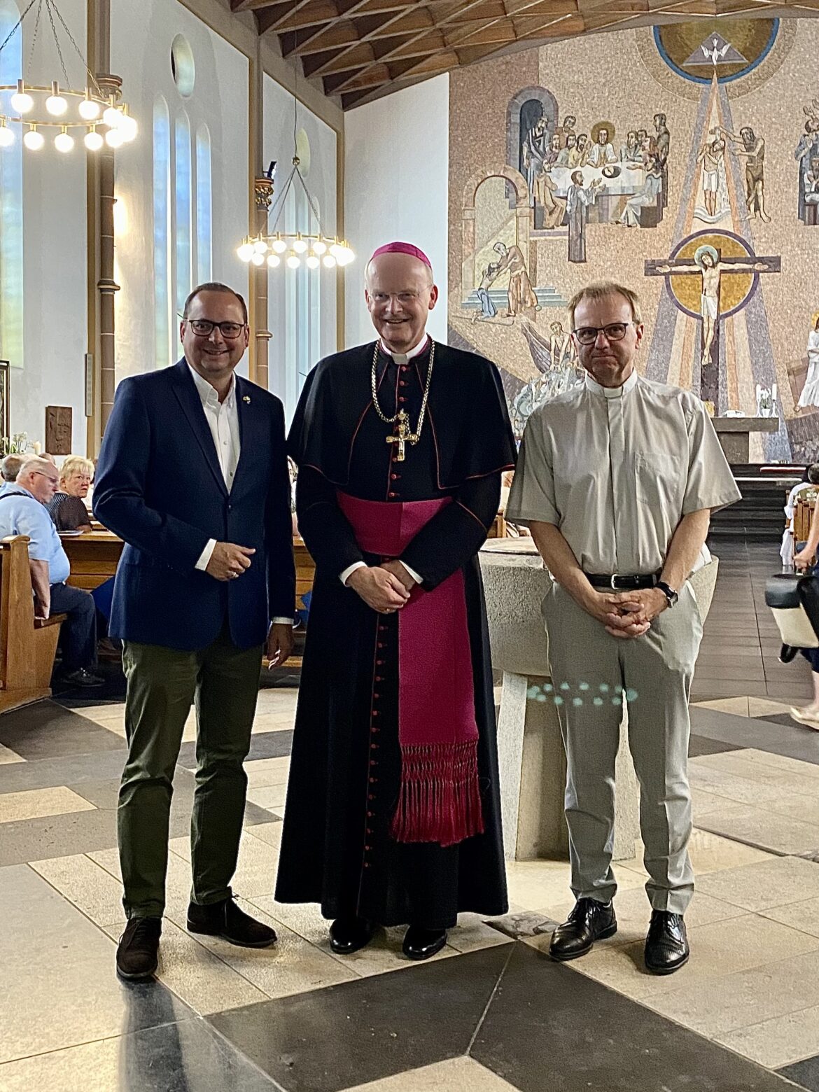 Essen‘s Oberbürgermeister, Bischof Franz-Josef Overbeck und Pfarrer Michael Dörnemann stehen vor dem Taufbecken der Kirche und lächeln in die Kamera.
