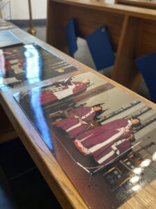 Auf einer Kirchenbank liegen mehrere Fotos hintereinander, die Szenen aus früheren Messen der Sankt Gertrud zeigen. Auf einem Foto sieht man mehrere Männer in weiß-lila Gewändern.