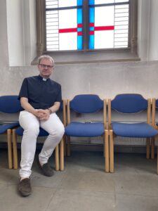 Michael Dörnemann sitzt auf einem Holzstuhl mit blauem Polster. Er befindet sich in einem Seitenraum der Sankt Gertrud Kirche in Essen, der hohe weiße Wände und eine blaue Decke hat.