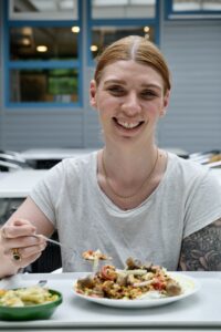 Julia Heinz sitzt in der Mensa mit ihrem Tablett vor sich und probiert lächelnd den Linsensalat