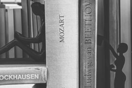 Das Bild zeigt eine Nahaufnahme eines Bücherregals in schwarz-weiß. Zwischen Büchern über bekannte europäische Komponisten wie Stockhausen, Mozart oder Beethoven sitzen und stehen zwei schattenhafte Frauenfiguren.