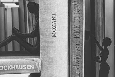 Das Bild zeigt eine Nahaufnahme eines Bücherregals in schwarz-weiß. Zwischen Büchern über bekannte europäische Komponisten wie Stockhausen, Mozart oder Beethoven sitzen und stehen zwei schattenhafte Frauenfiguren.