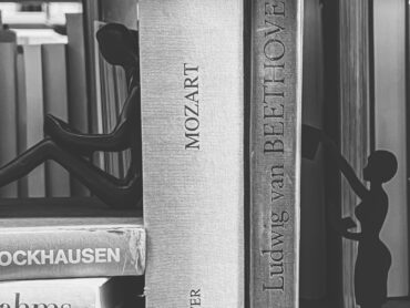 Das Bild zeigt eine Nahaufnahme eines Bücherregals in schwarz-weiß. Zwischen Büchern über bekannte europäische Komponisten wie Stockhausen, Mozart oder Beethoven sitzen und stehen zwei schattenhafte Frauenfiguren.