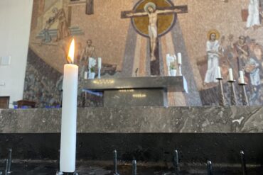 Eine einzige Kerze steht vor dem Altar der Sankt Gertrud Kirche in Essen. Im Hintergrund ist das große braune Wandmosaik zu erkennen, dass Jesus am Kreuz zeigt.