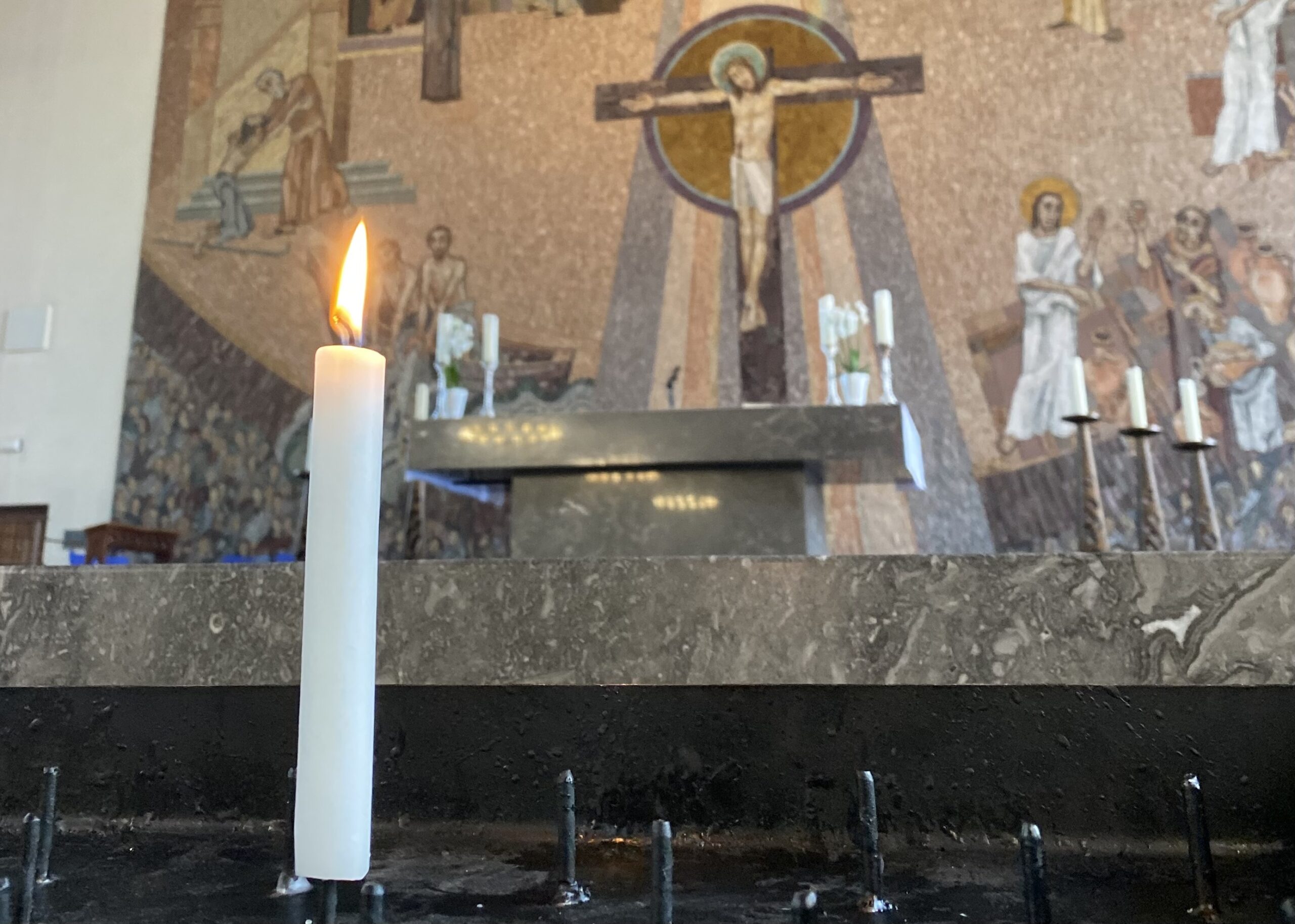 Eine einzige Kerze steht vor dem Altar der Sankt Gertrud Kirche in Essen. Im Hintergrund ist das große braune Wandmosaik zu erkennen, dass Jesus am Kreuz zeigt.