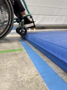 Ein Rollstuhl vor einer Turnmatte. Die kleinen Vorderräder werden auf der Matte abgesetzt.