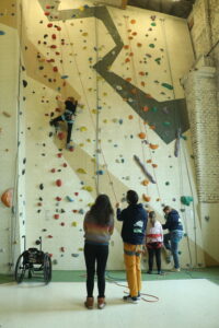 Zwei Menschen stehen mittig im Bild vor einer Kletterwand und schauen nach oben. Auf halber Höhe der Wand befindet sich links im Bild eine kletternde Person. Unten links an der Wand steht ein Rollstuhl.