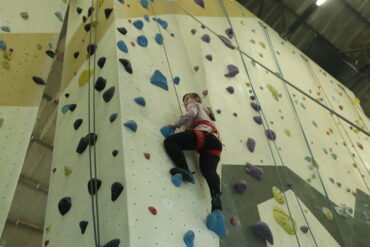 Blick auf eine Kletterwand an der eine Frau auf halber Höhe nach oben klettert.