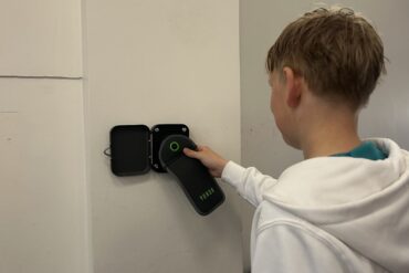 Ein Schüler in einem weißen Kapuzenpullover steht vor einer Magnetstation im Gymnasium Schaurtestr in Köln. Er hält seine Handytasche an den Magneten, um sie zu öffnen.