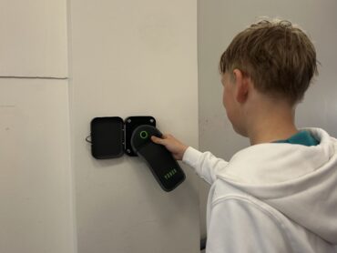 Ein Schüler in einem weißen Kapuzenpullover steht vor einer Magnetstation im Gymnasium Schaurtestr in Köln. Er hält seine Handytasche an den Magneten, um sie zu öffnen.