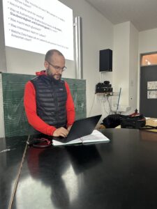 Jens Wenzel, Lehrer und Medienkoordinator am Gymnasium Schaurtestraße in Köln, arbeitet an einem Laptop vor einer grünen Schultafel. Hinter ihm ist eine Präsentation an die Wand projiziert.